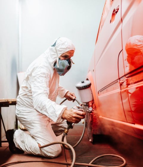 mechanic male worker painting a car in a special painting box, wearing a white costume and protection gear